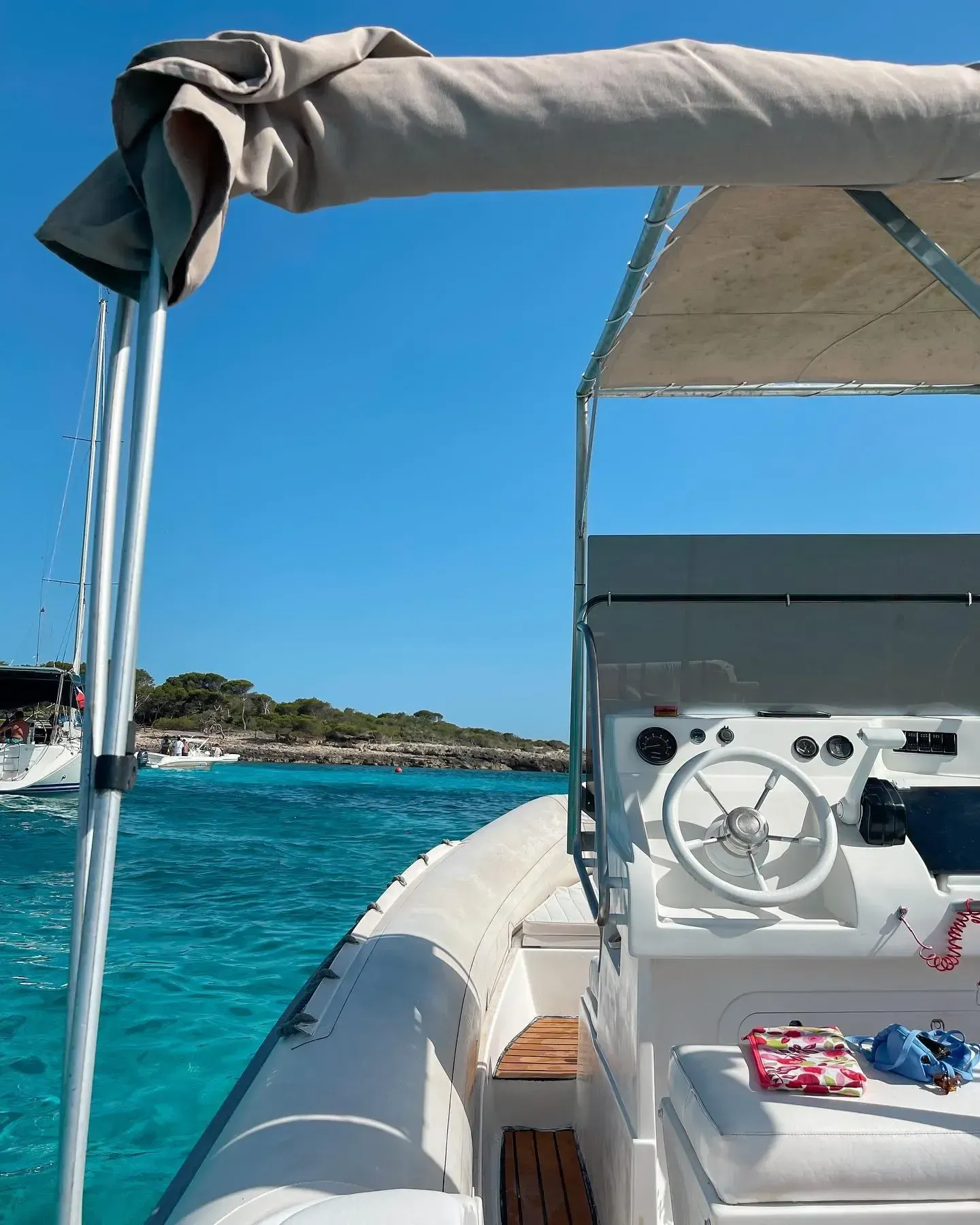 Vista desde el timón de un barco blanco, contemplando las aguas turquesas y una lejana costa rocosa bajo un cielo azul.