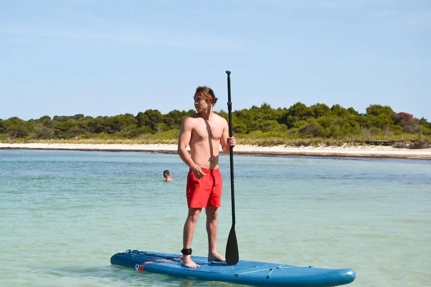 Una persona con bañador rojo está de pie sobre una tabla de paddle surf azul en aguas claras y poco profundas, cerca de una playa de arena y árboles verdes.