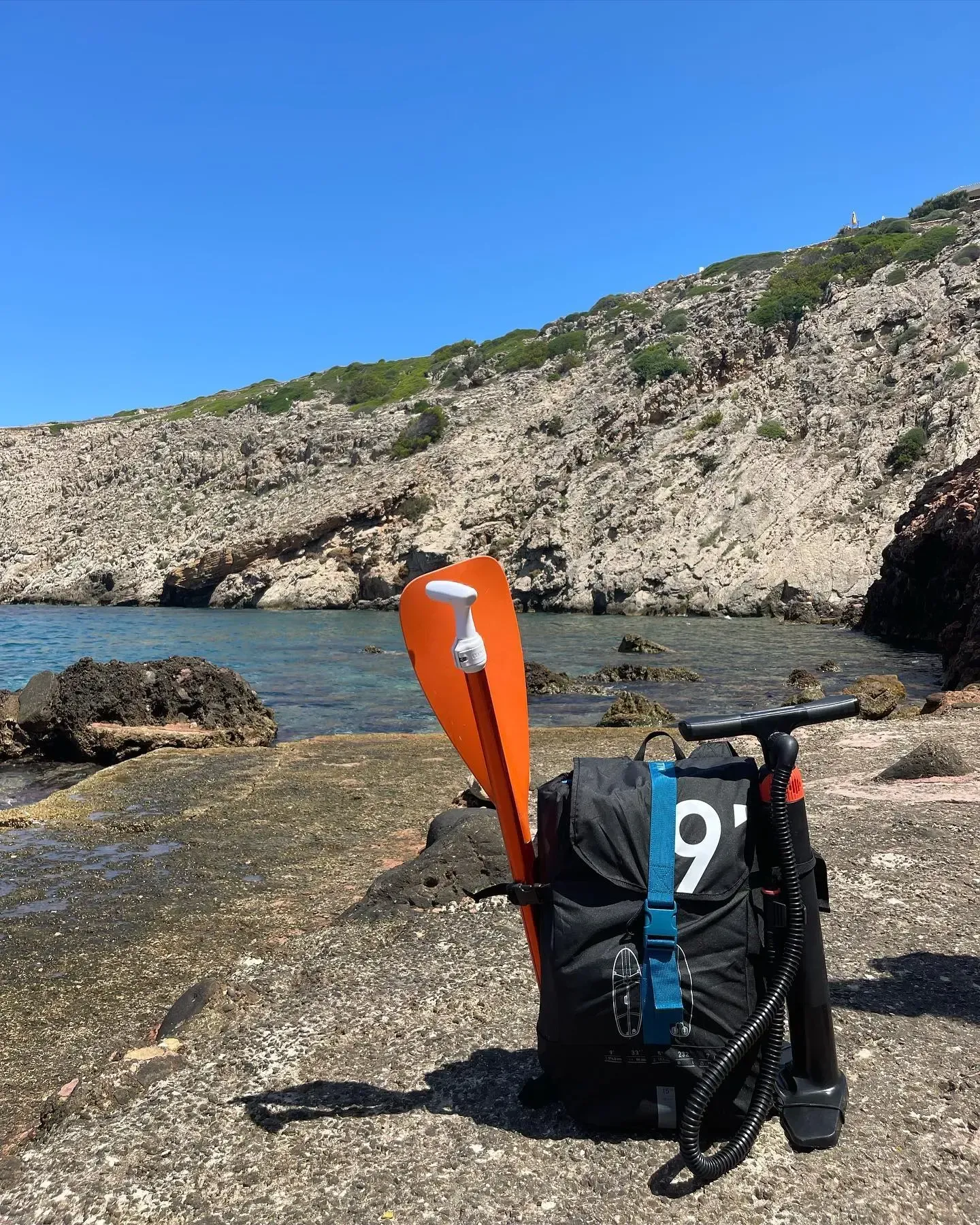 Una mochila para tabla de paddle surf y un remo de color naranja brillante reposan en una orilla rocosa frente a una soleada bahía costera.