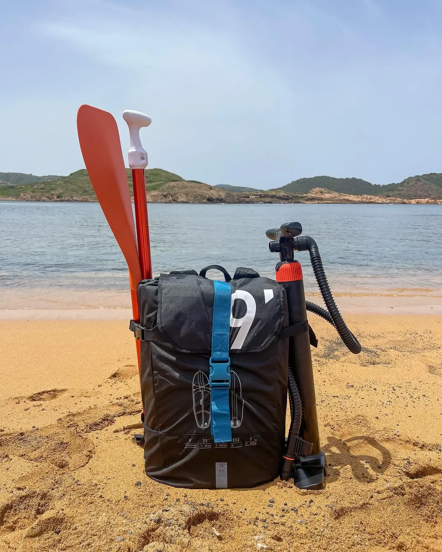 Una mochila negra con un remo naranja y una bomba de aire se encuentra en una playa de arena, con un océano en calma y colinas al fondo.