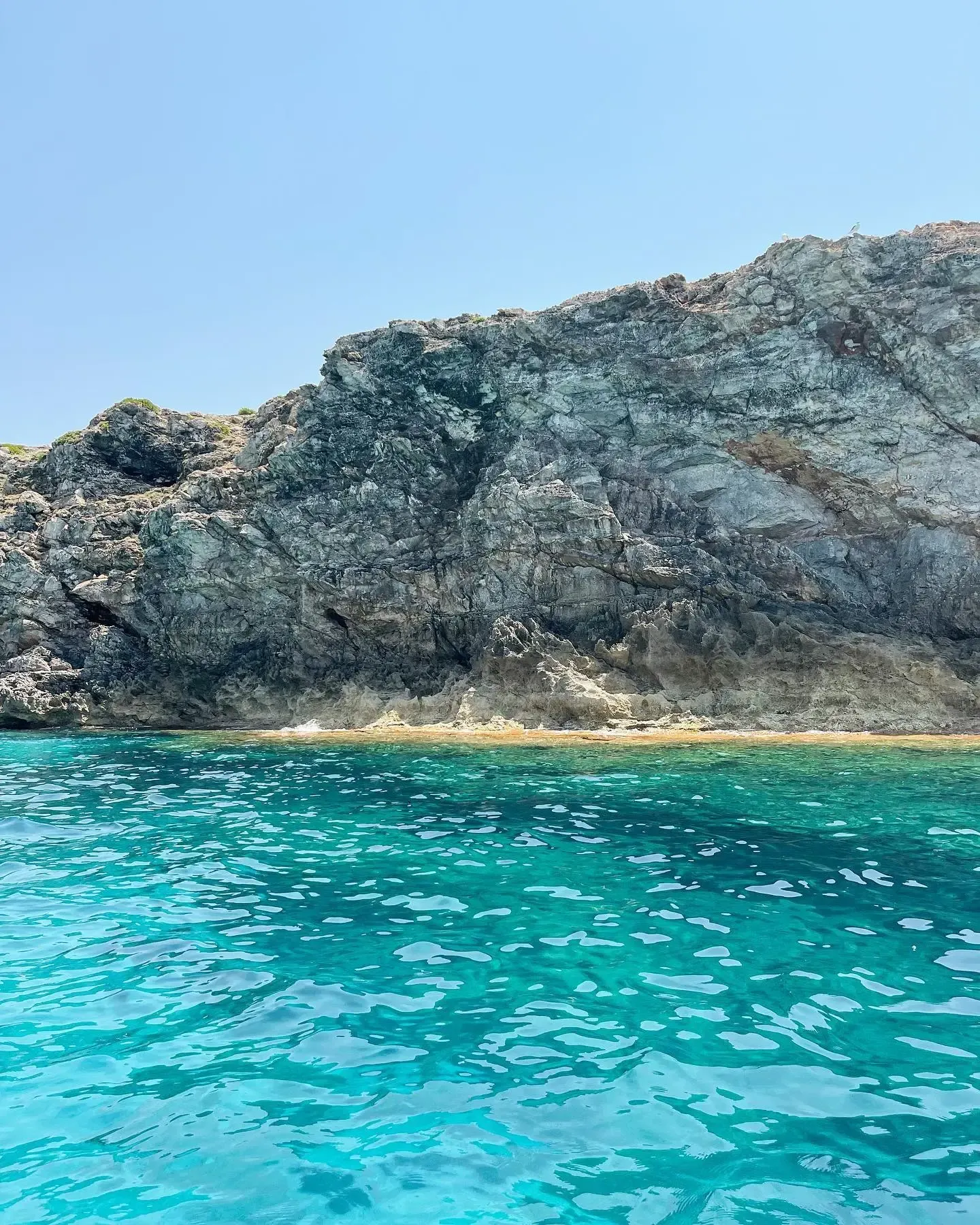 En primer plano, aguas cristalinas de color turquesa se encuentran con un escarpado acantilado rocoso bajo un cielo azul brillante y sin nubes.