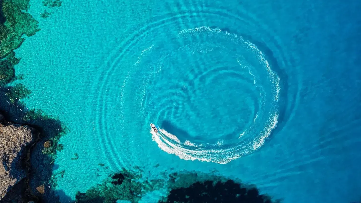 Vista aérea de una pequeña embarcación dejando una estela circular en aguas de color turquesa brillante cerca de una costa rocosa.