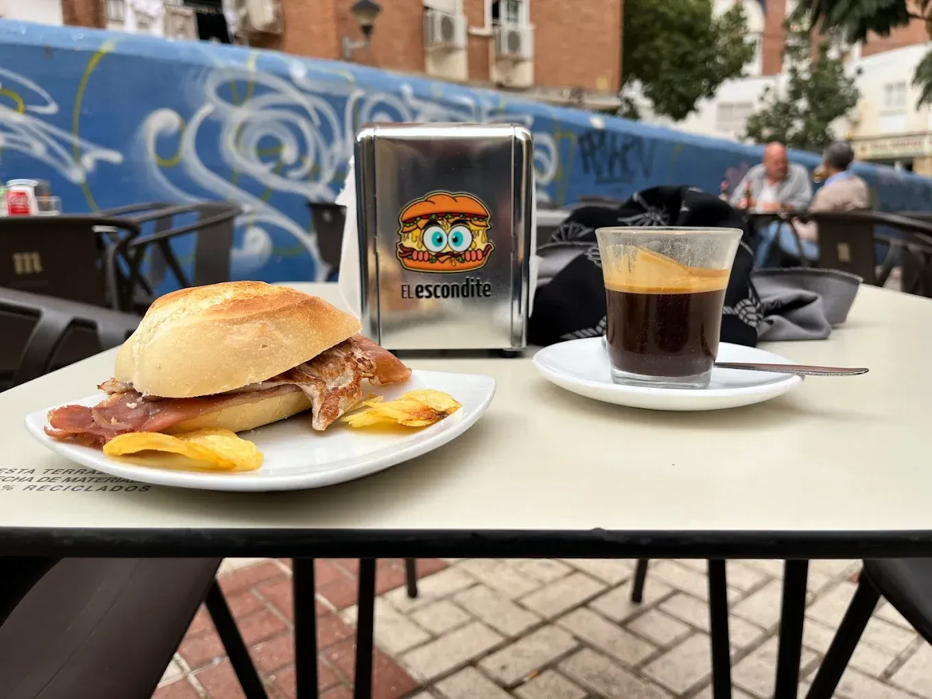 Un sándwich y un café en la mesa de una terraza. El sándwich lleva carne y patatas fritas.