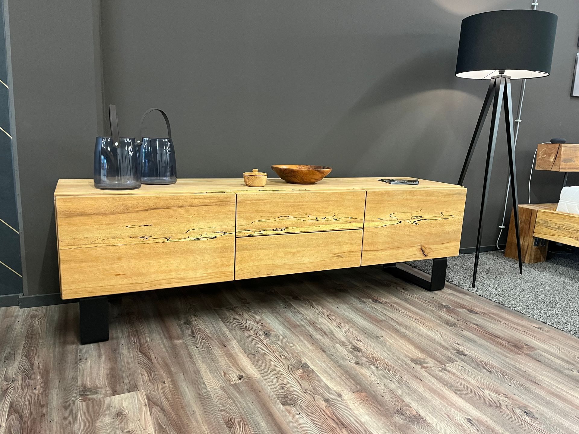 Holzschrank mit schwarzen Metallbeinen, darauf Vasen und eine Schale; daneben eine Lampe.