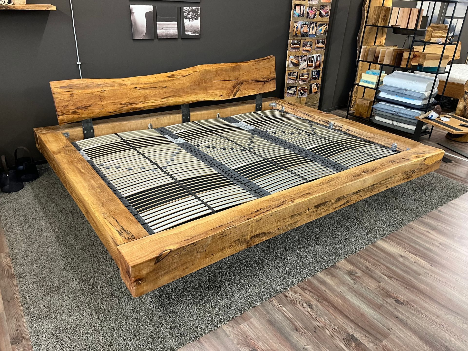 Holzplattformbettgestell mit rustikalem Kopfteil auf einem grauen Teppich in einem Ausstellungsraum.