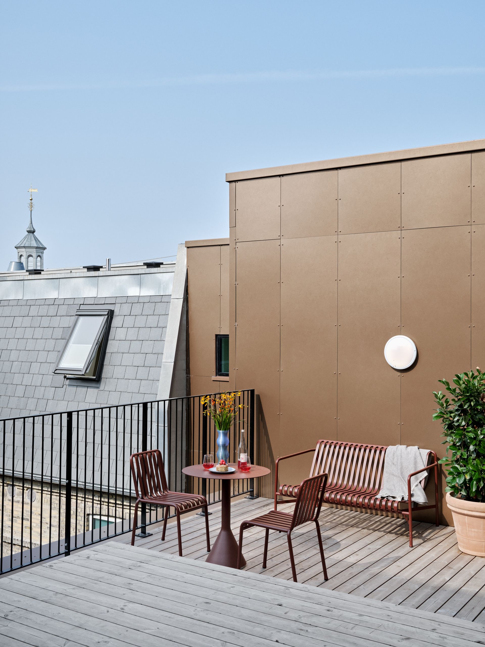 Rooftop patio with brown furniture, small table, and potted plant. City buildings in the background.