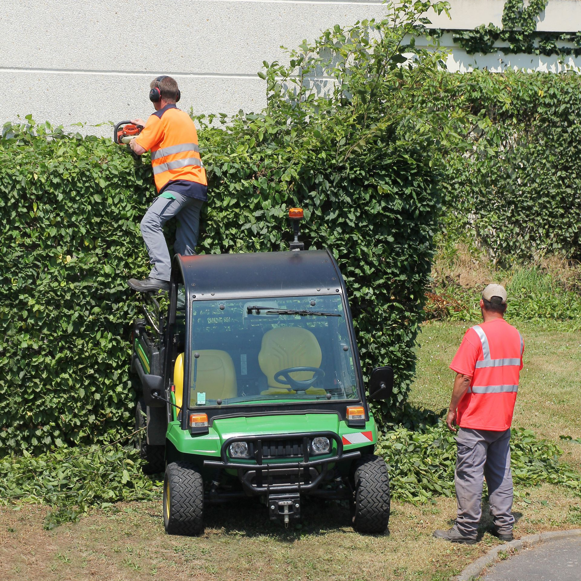 Un homme taille une haute haie, debout sur un petit véhicule utilitaire. Un autre homme observe.