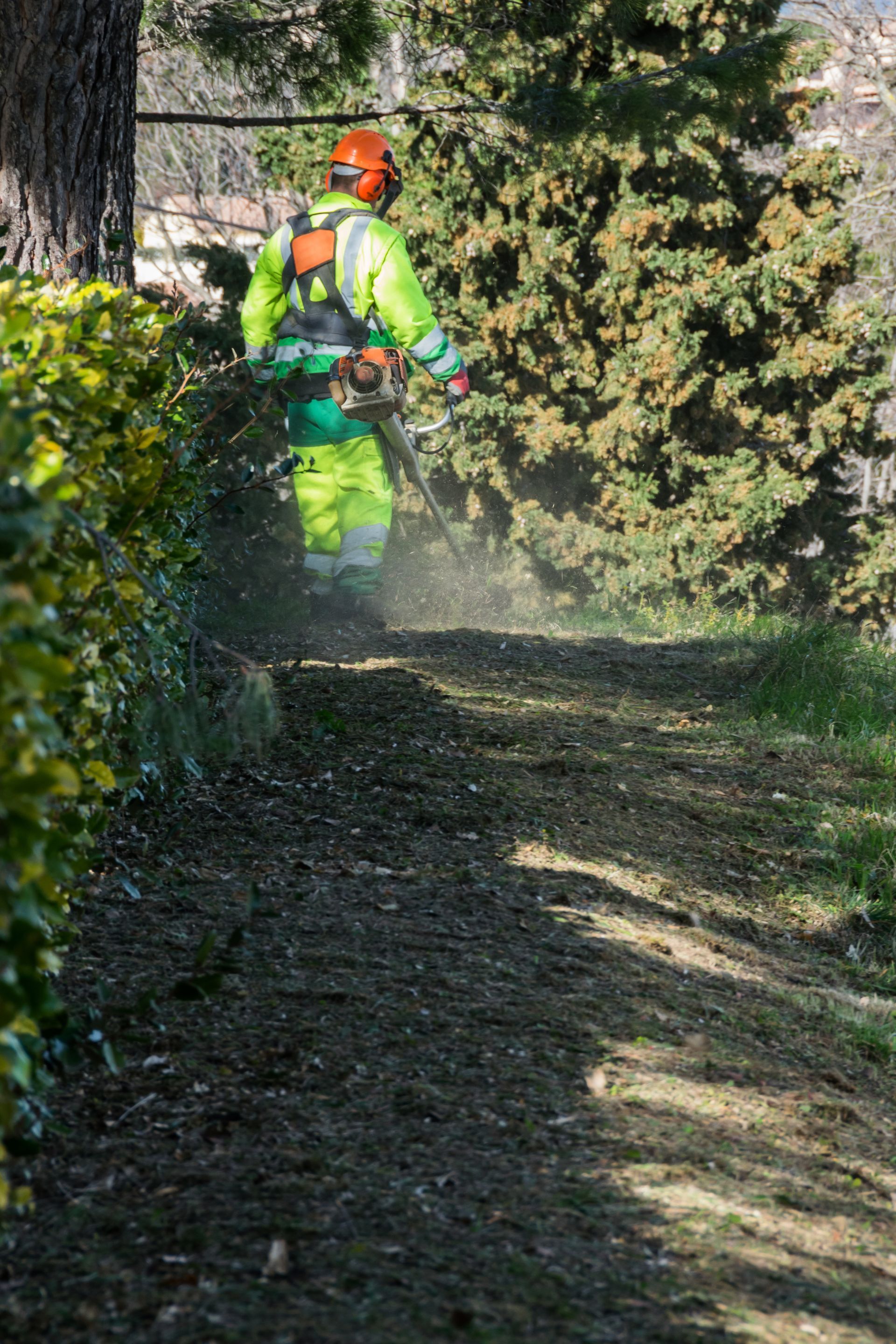 Une personne portant un équipement de sécurité utilise une débroussailleuse sur un chemin de terre bordé de végétation.