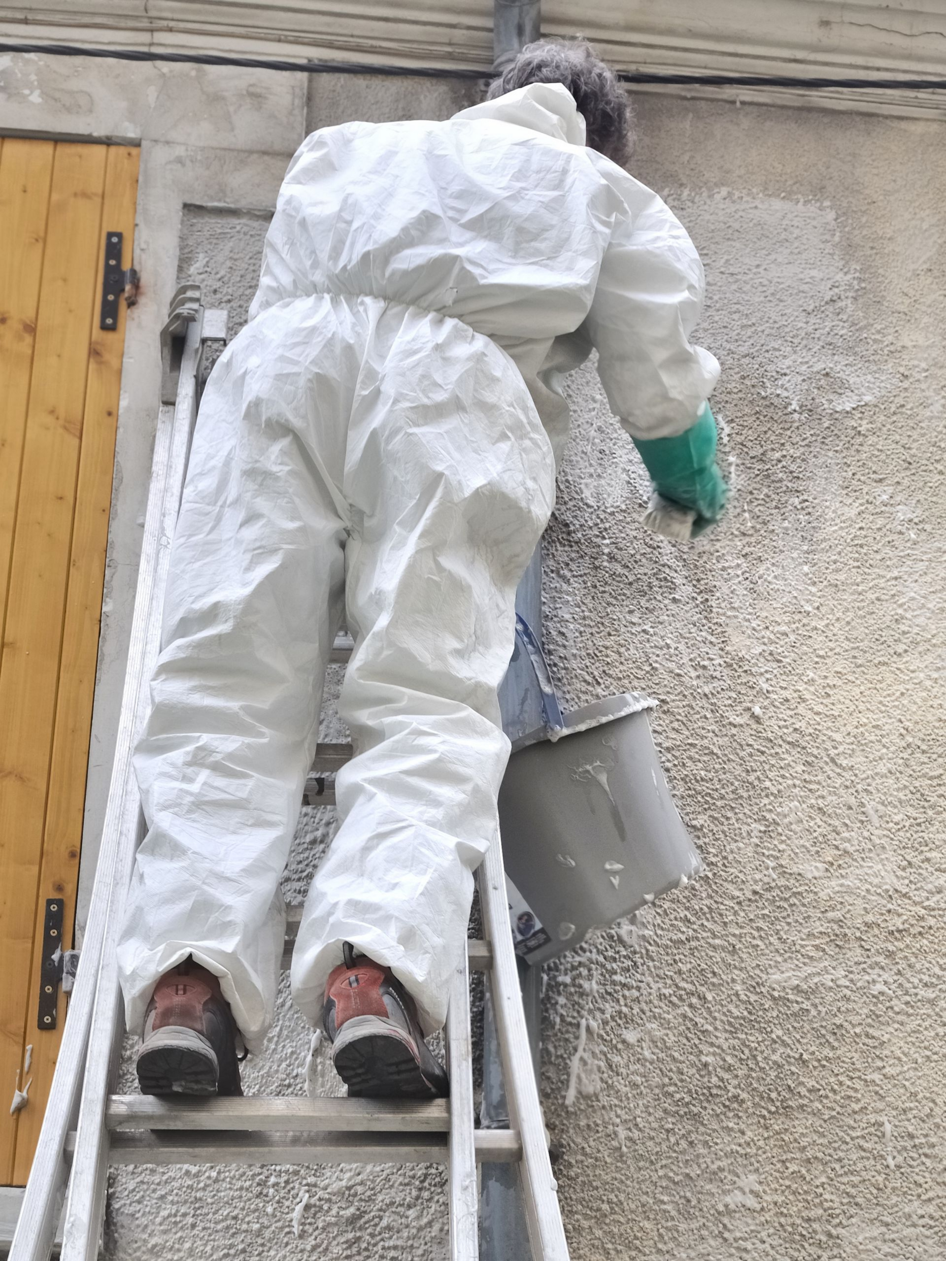 Personne en combinaison de protection blanche sur une échelle appliquant une substance sur un mur, portant un seau.
