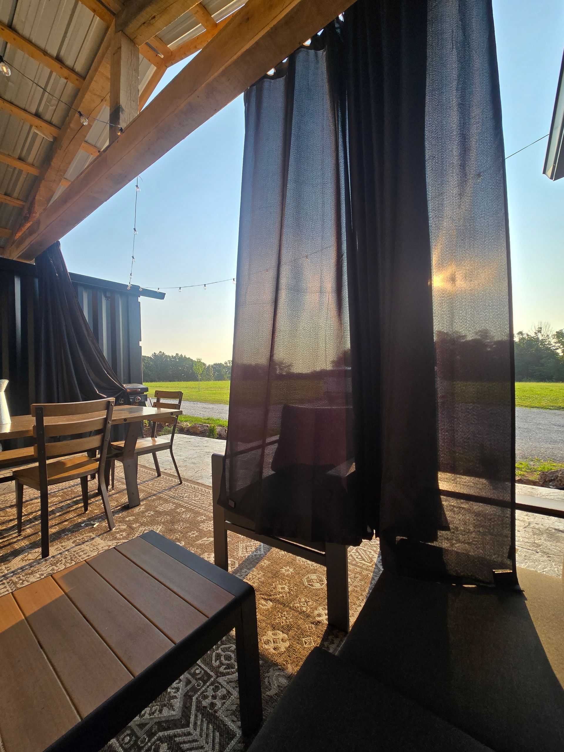 Outdoor patio with black curtains, wooden furniture, and sunlight over a grassy view beyond the railing