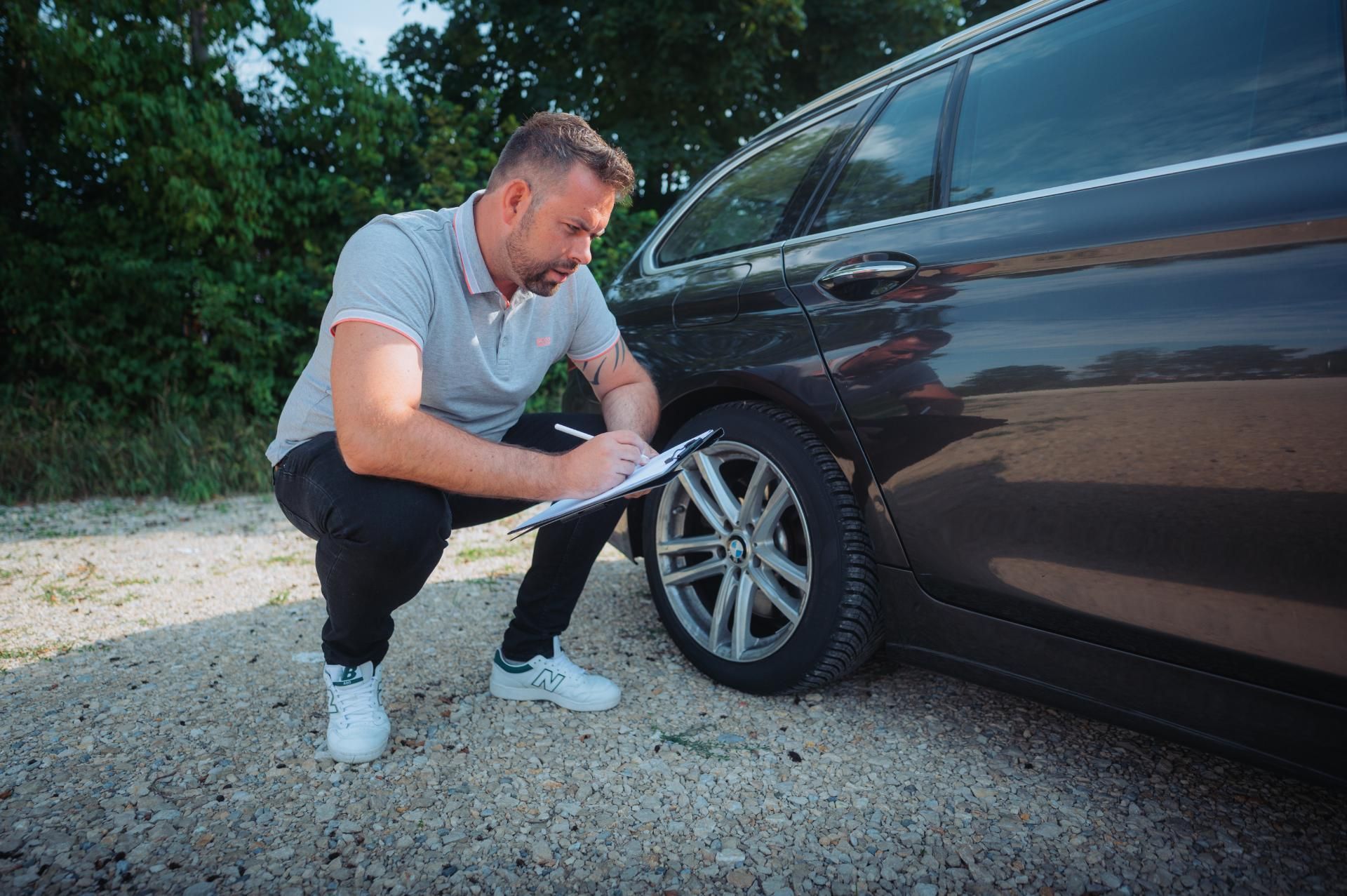 ein Mann hockt neben einem schwarzen Auto und notiert sich etwas auf einem Klemmbrett