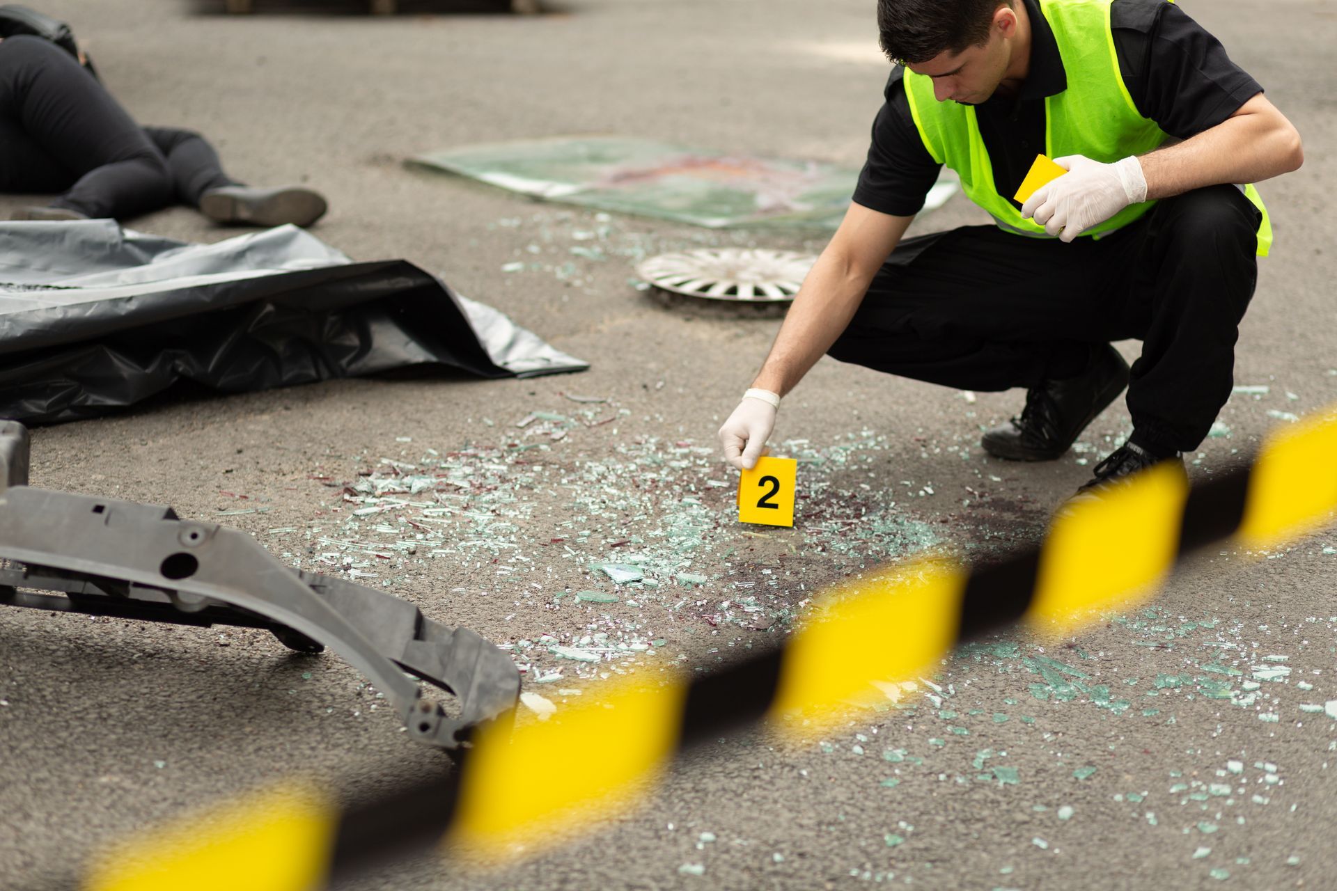 Tatort: Ermittler in Warnweste untersucht Glasscherben auf Asphalt, Leiche bedeckt, Trümmer, gelbe Spurenmarkierung.