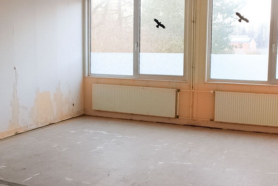 Leerer Raum mit großen Fenstern, Heizkörpern und Wasserschäden an den Wänden.