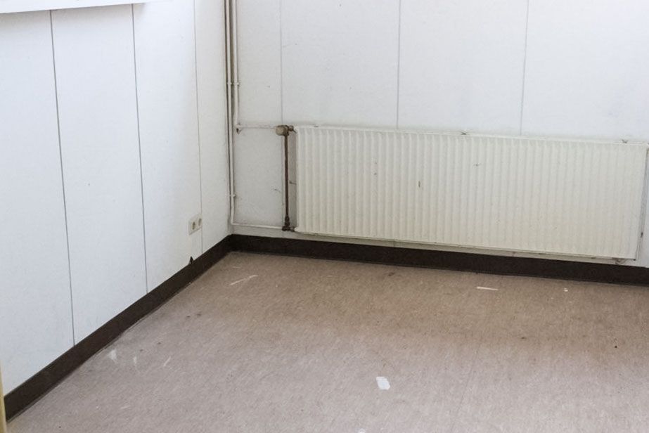 Leerer Raum mit weißen Paneelwänden, braunem Boden und Heizkörper.