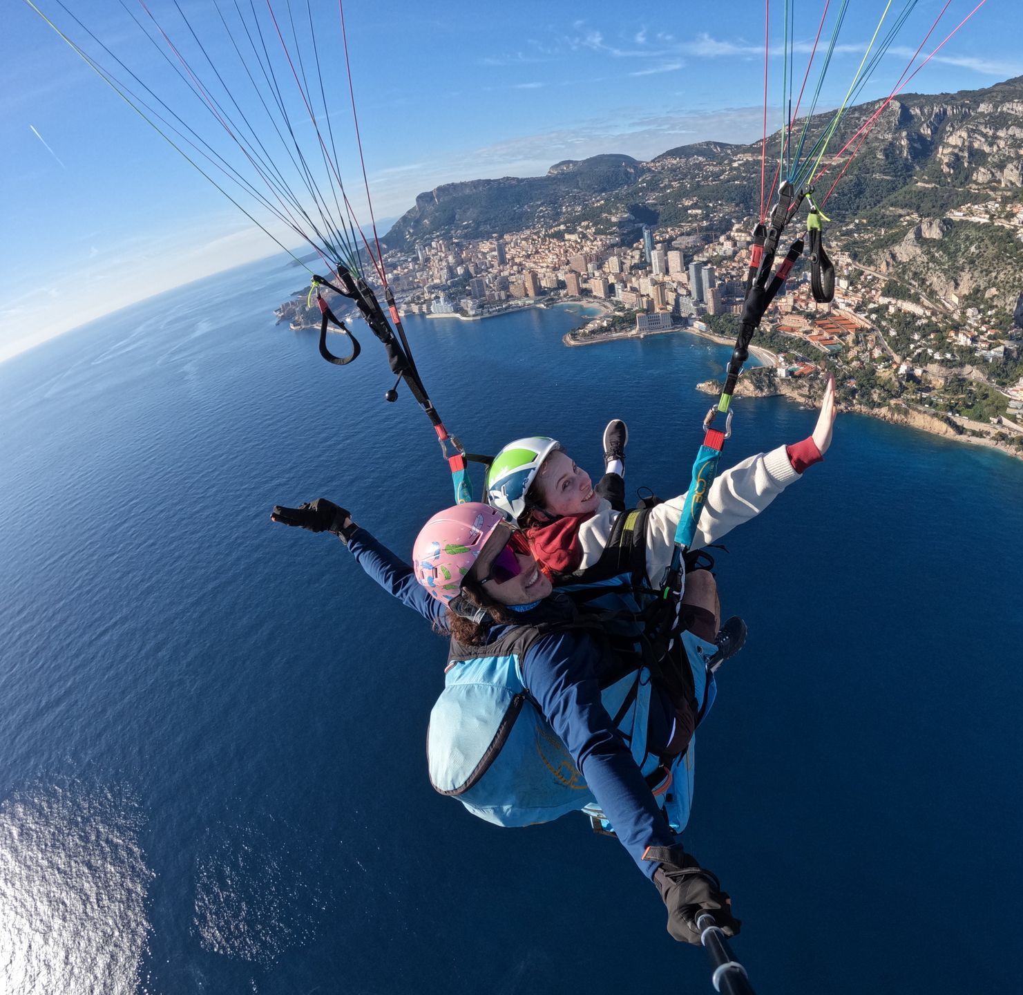 Two people paragliding over a coastal city, blue sea, sky. Smiles, colorful canopy.