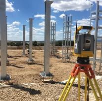 Un instrumento de topografía sobre un trípode, inspeccionando un sitio de construcción con pilares de metal.