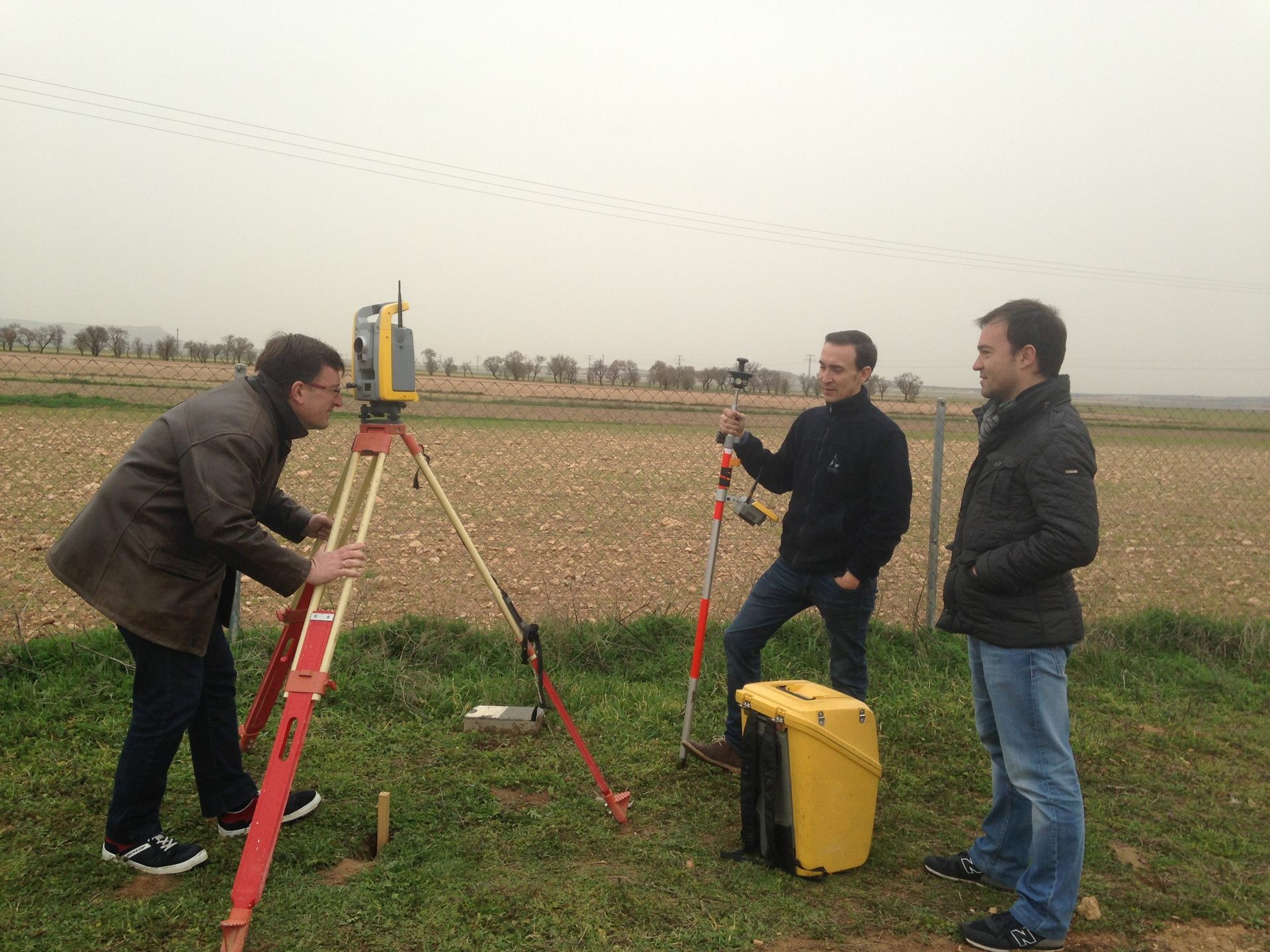 Dispositivo de medición amarillo sobre trípode, vías del tren, tren al fondo, paredes de hormigón, día soleado.