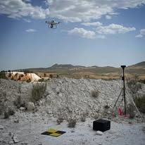 Dispositivo de medición amarillo sobre trípode, vías del tren, tren al fondo, paredes de hormigón, día soleado.