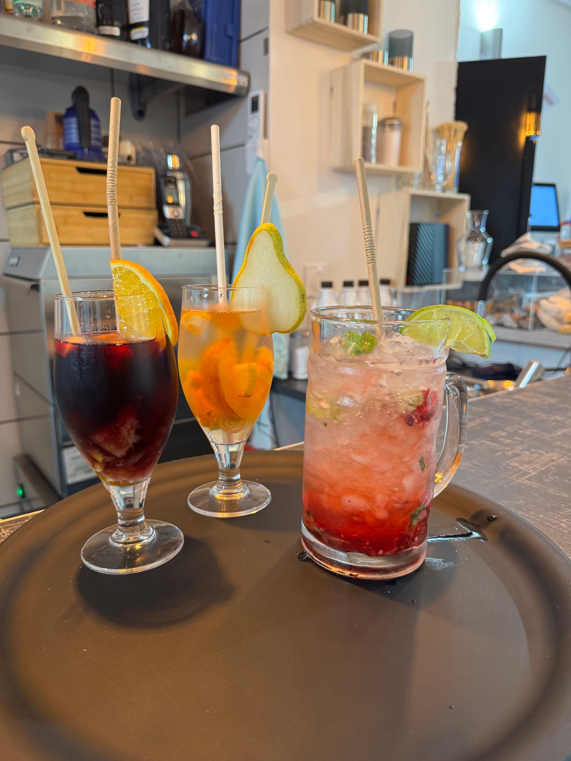 Trois cocktails colorés avec des garnitures de fruits sur un plateau dans un décor de café.