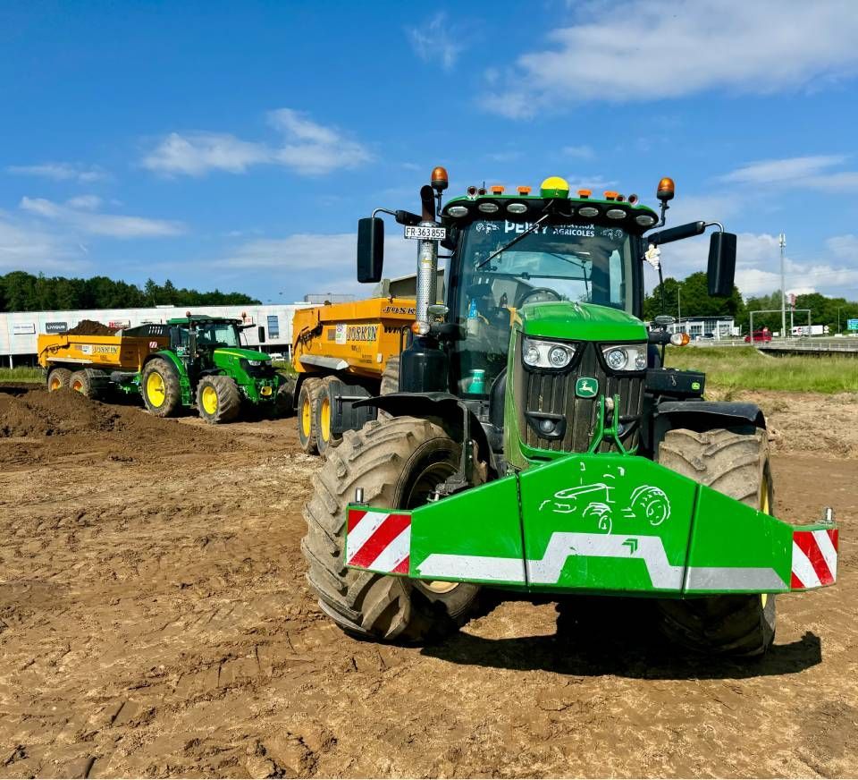 Tracteur John Deere vert sur un chantier de construction, tirant deux remorques sous un ciel bleu.