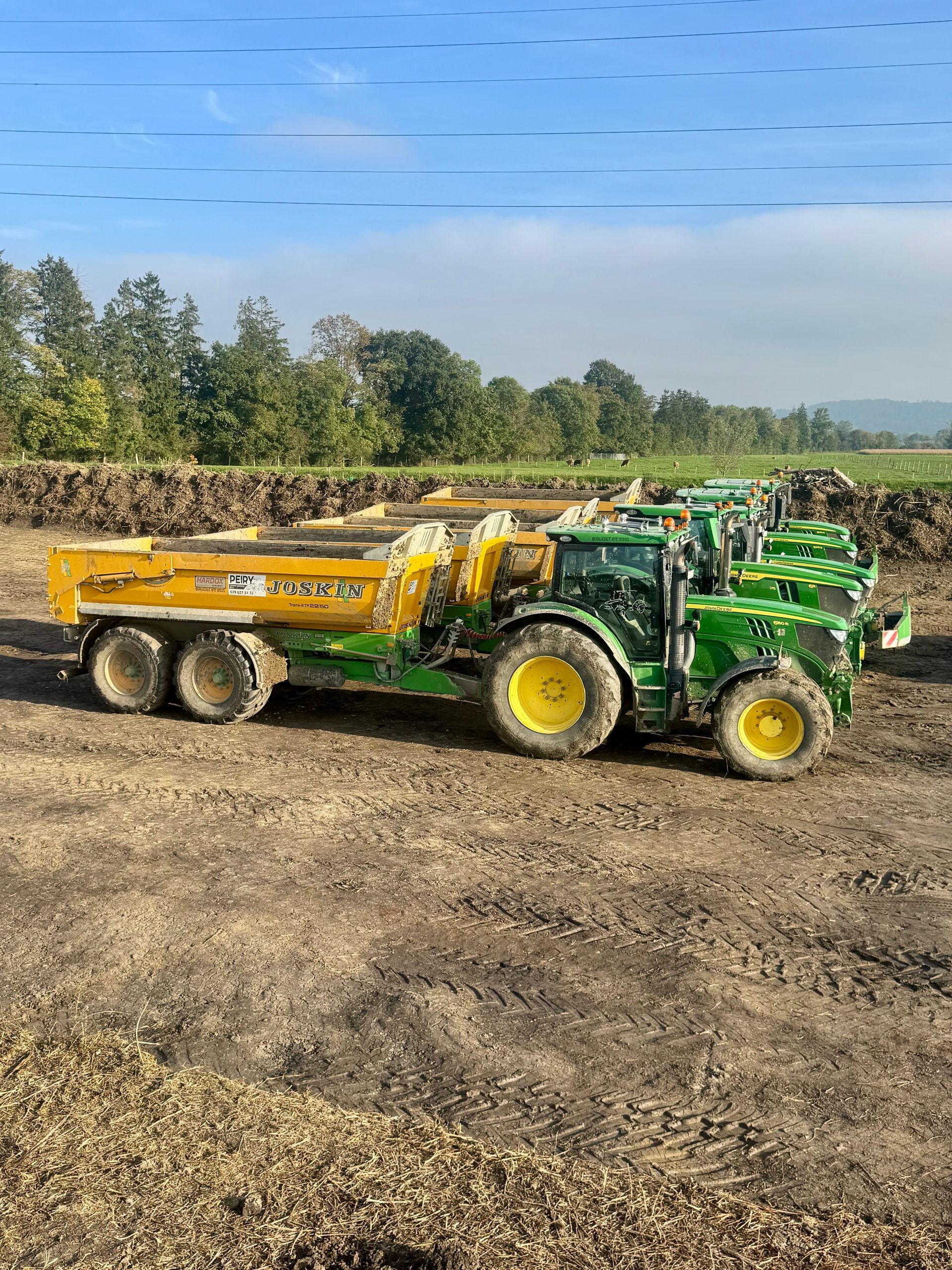 Plusieurs tracteurs John Deere avec des remorques basculantes jaunes sur un champ boueux sous un ciel bleu.
