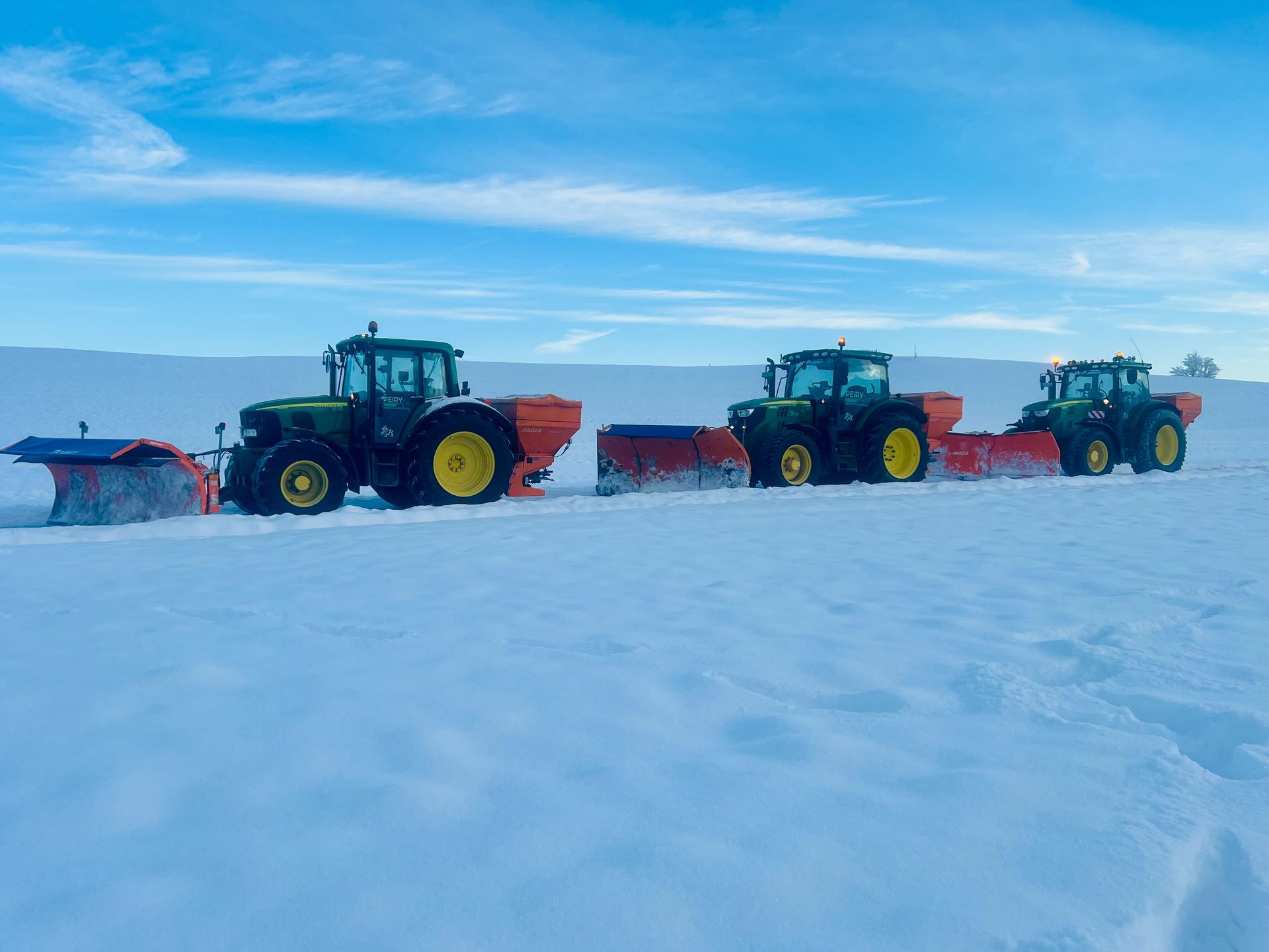 Trois tracteurs John Deere équipés de chasse-neige sur un champ enneigé sous un ciel bleu.