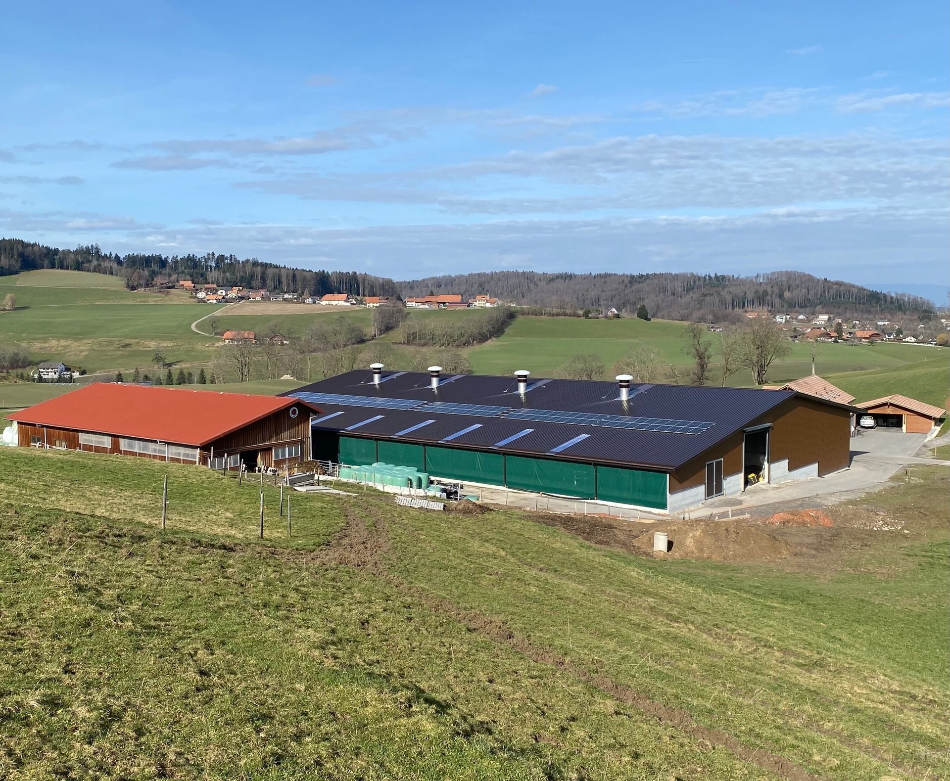 Des bâtiments de ferme aux toits rouges, verts et bruns se dressent sur un flanc de colline herbeux sous un ciel dégagé.