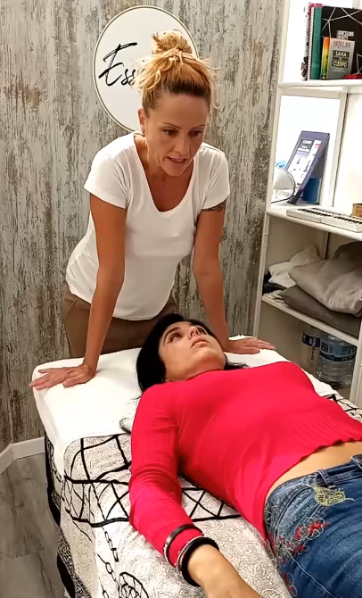 Dona amb camisa blanca inclinada sobre un pacient amb samarreta vermella sobre una taula de massatge. Sala de tractament.