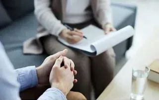 Terapeuta tomando notas durante una sesión; las manos del paciente entrelazadas en el regazo, expresiones neutrales.