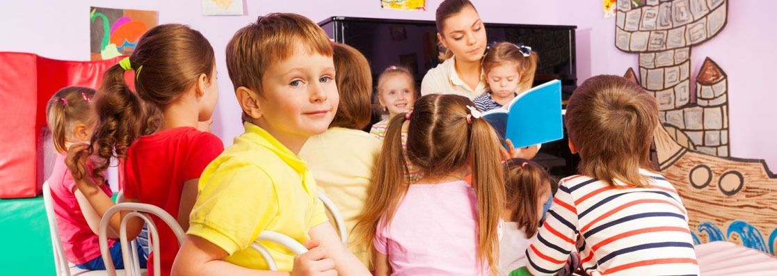 Un grupo de niños está sentado en un aula escuchando a un profesor leer un libro.
