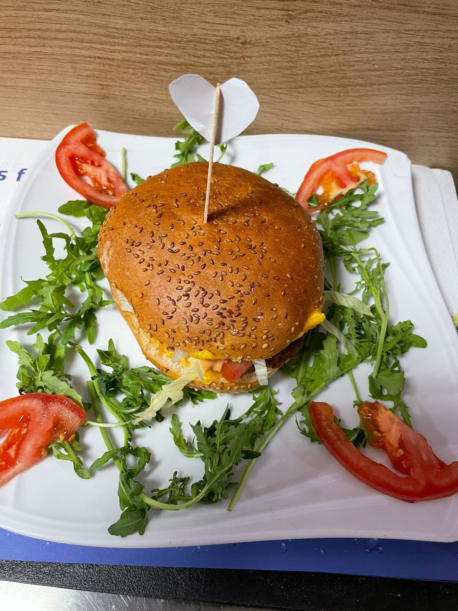 Ein Burger auf einem weissen Teller, gekrönt mit einem herzförmigen Zahnstocher, garniert mit Rucola und Tomatenscheiben.