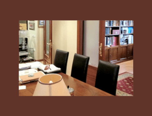 Mesa de oficina de madera con tres sillas negras, una lámpara y estanterías visibles al fondo.