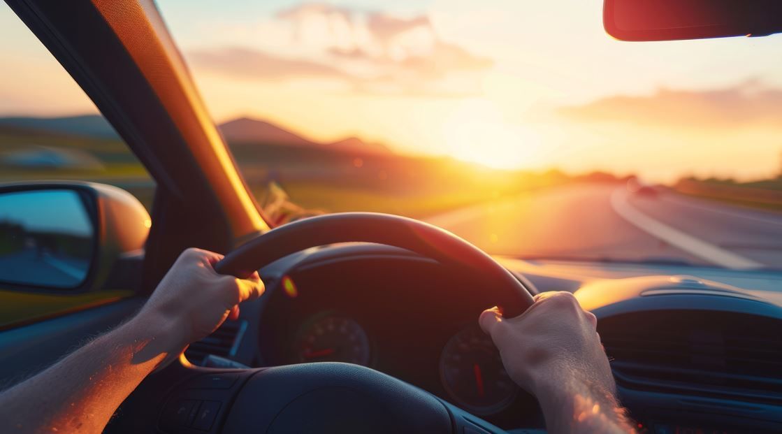 Manos agarrando un volante, conduciendo hacia una puesta de sol en una autopista.