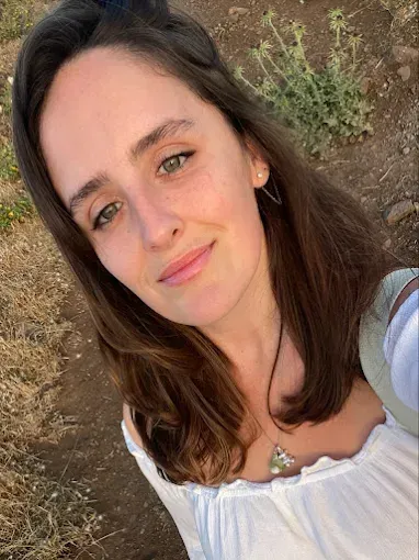 Mujer con blusa blanca sonríe afuera; cabello castaño, ojos verdes, entorno natural.