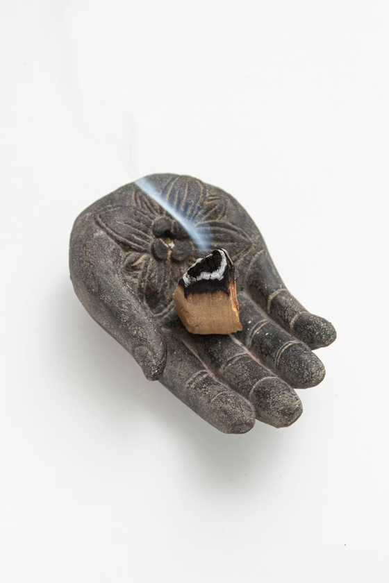 Porta incienso oscuro con forma de mano, que sostiene un cubo de incienso encendido del que se elevan volutas de humo blanco.