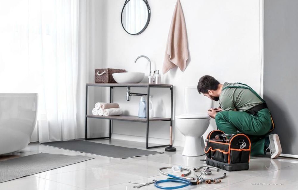 Ein Fachmann in Arbeitskleidung kniet auf dem Badezimmerboden, um eine weiße Toilette zu reparieren; Werkzeug liegt in der Nähe.