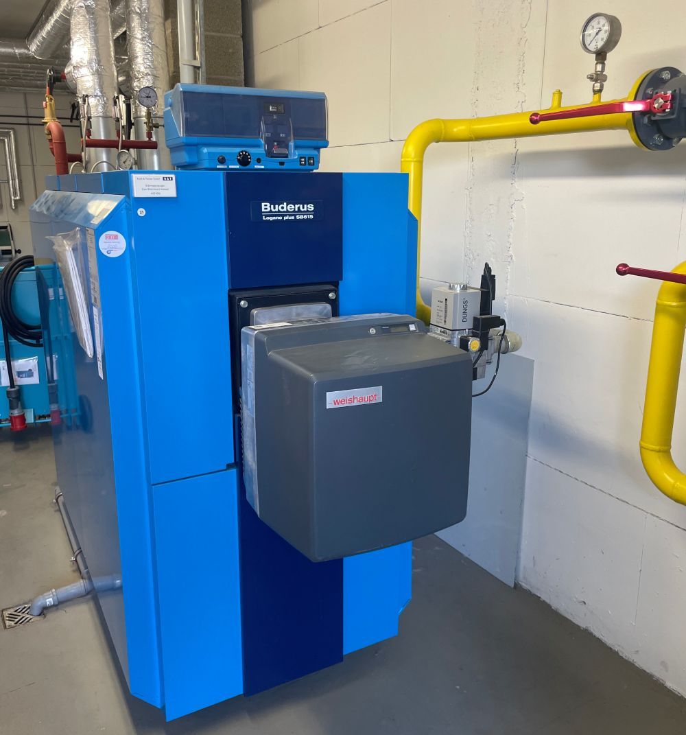 In einem Technikraum steht ein blauer Buderus-Industriekessel, an dessen Wand gelbe Gasleitungen verlaufen.