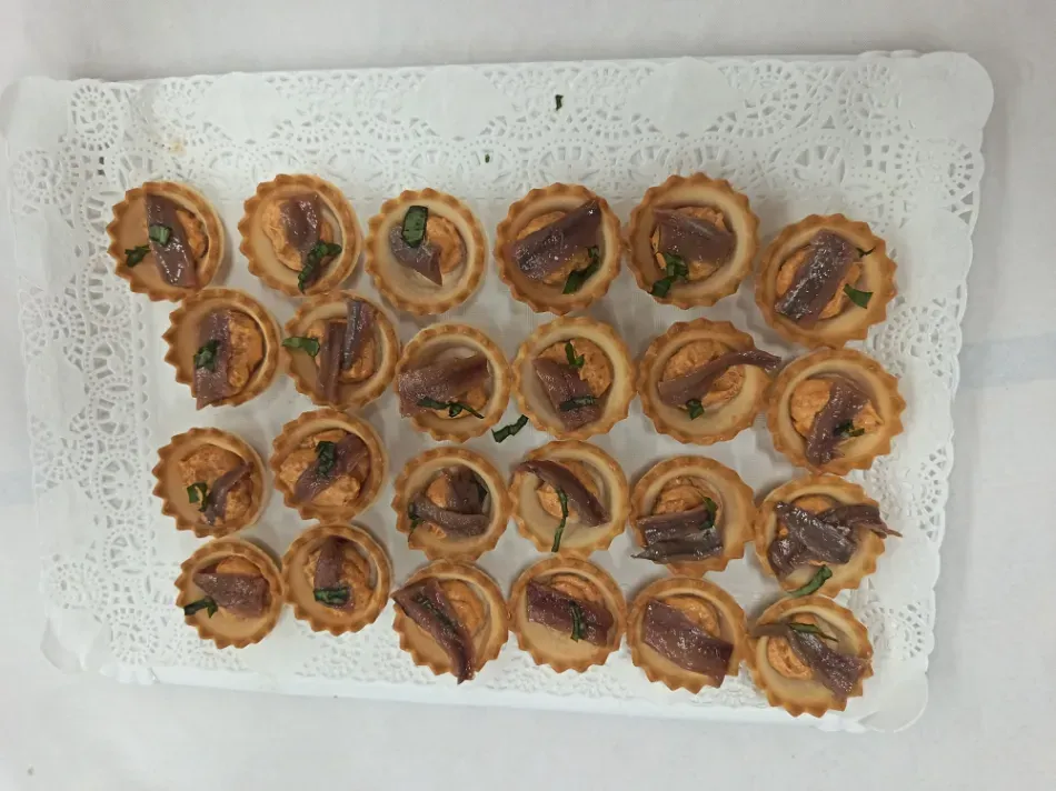 Pequeños pastelitos dispuestos en una bandeja cubierta con un mantelito de encaje blanco, algunos adornados con una guarnición verde.