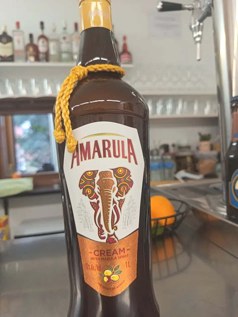 Botella de licor de crema de marula con etiqueta amarilla de cuerda en el cuello, sobre la barra de un bar con cristalería detrás