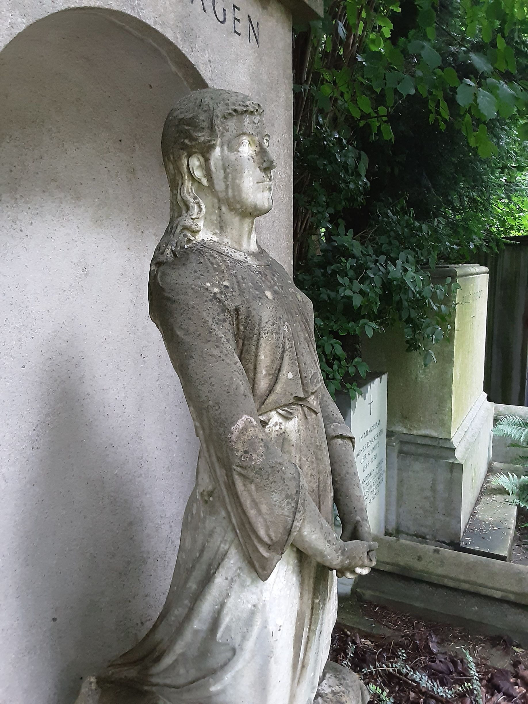 Steinstatue einer Frau in einem wallenden Gewand, die auf einen Friedhof blickt.