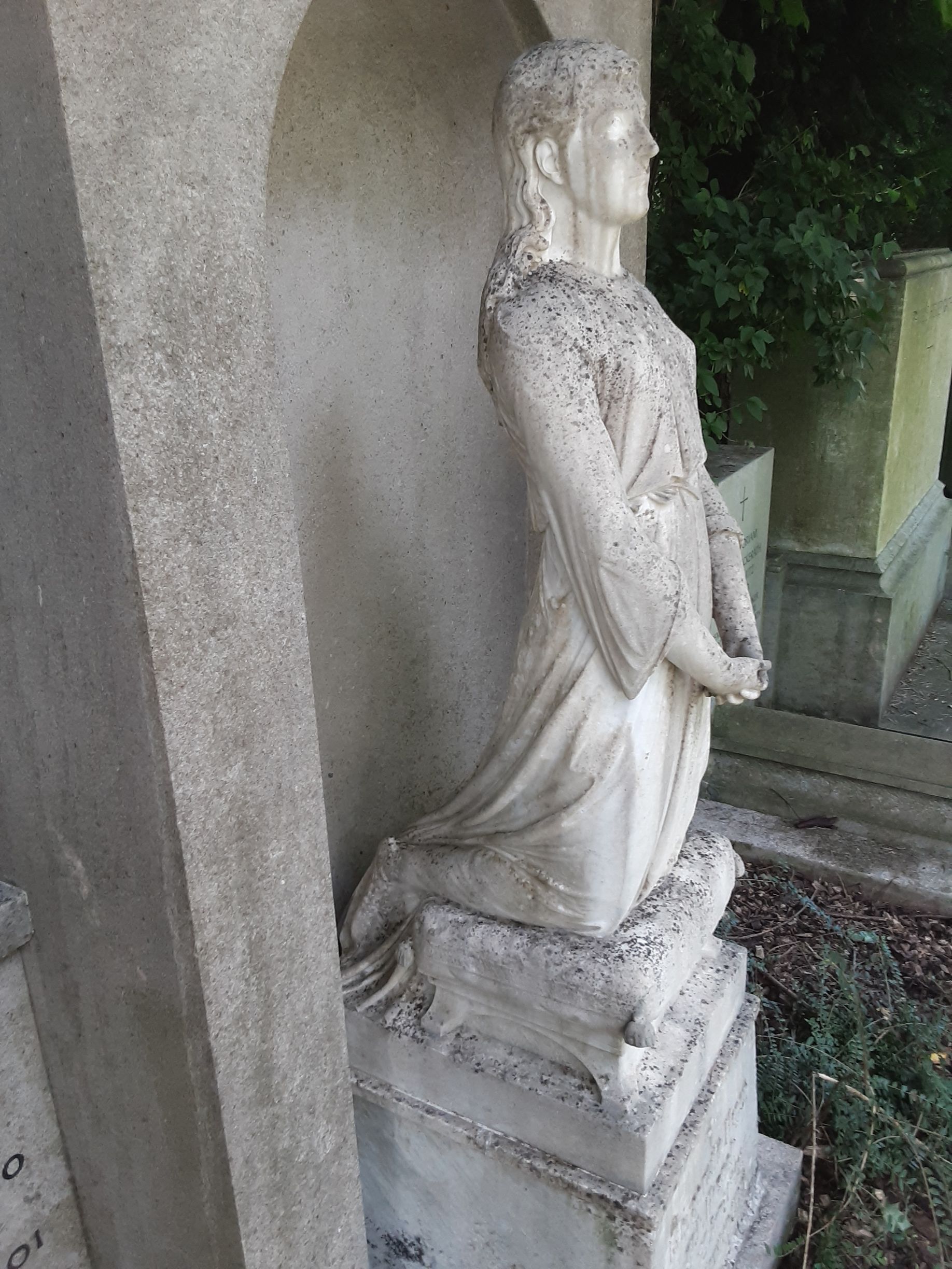 Statue einer knienden Figur in einer Nische. Heller Stein mit verwitterter Textur, in einer Friedhofsumgebung.
