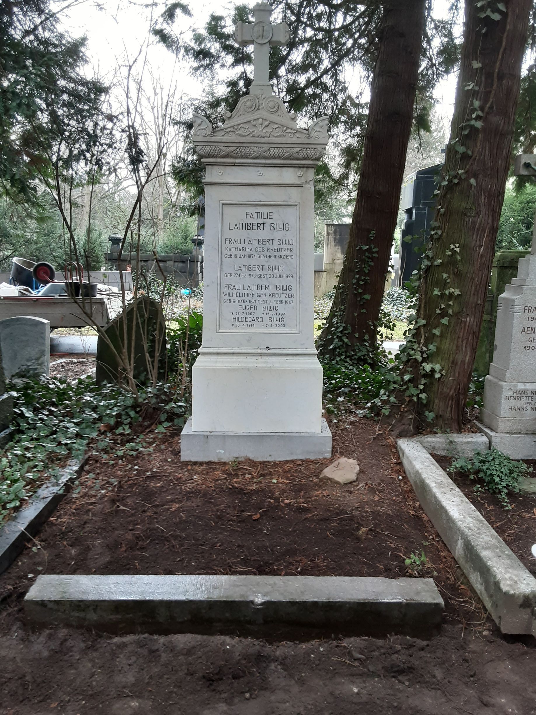 Weißer Grabstein mit einem Kreuz darauf, in einer Friedhofsumgebung.