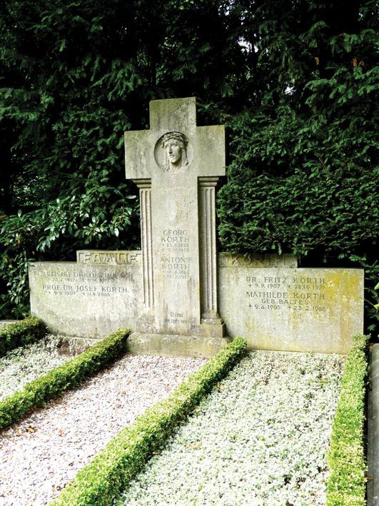 Steingrab mit kreuzförmigem Denkmal, Porträt und Inschriften. Grüne Sträucher umrahmen die Grabstätte.
