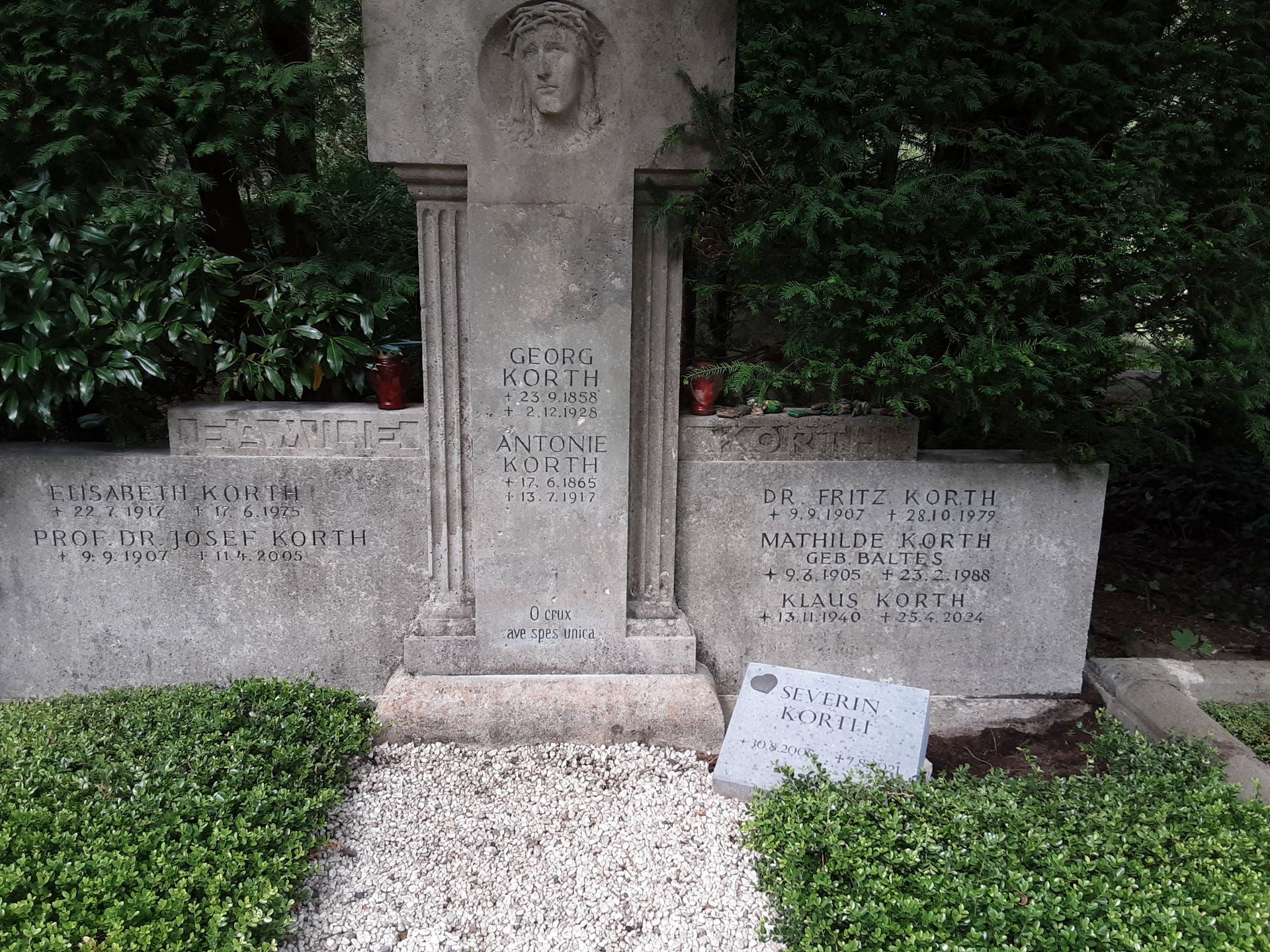 Grabmal aus grauem Stein mit eingemeißeltem Gesicht, Kreuz und Inschriften, im Hintergrund Büsche.