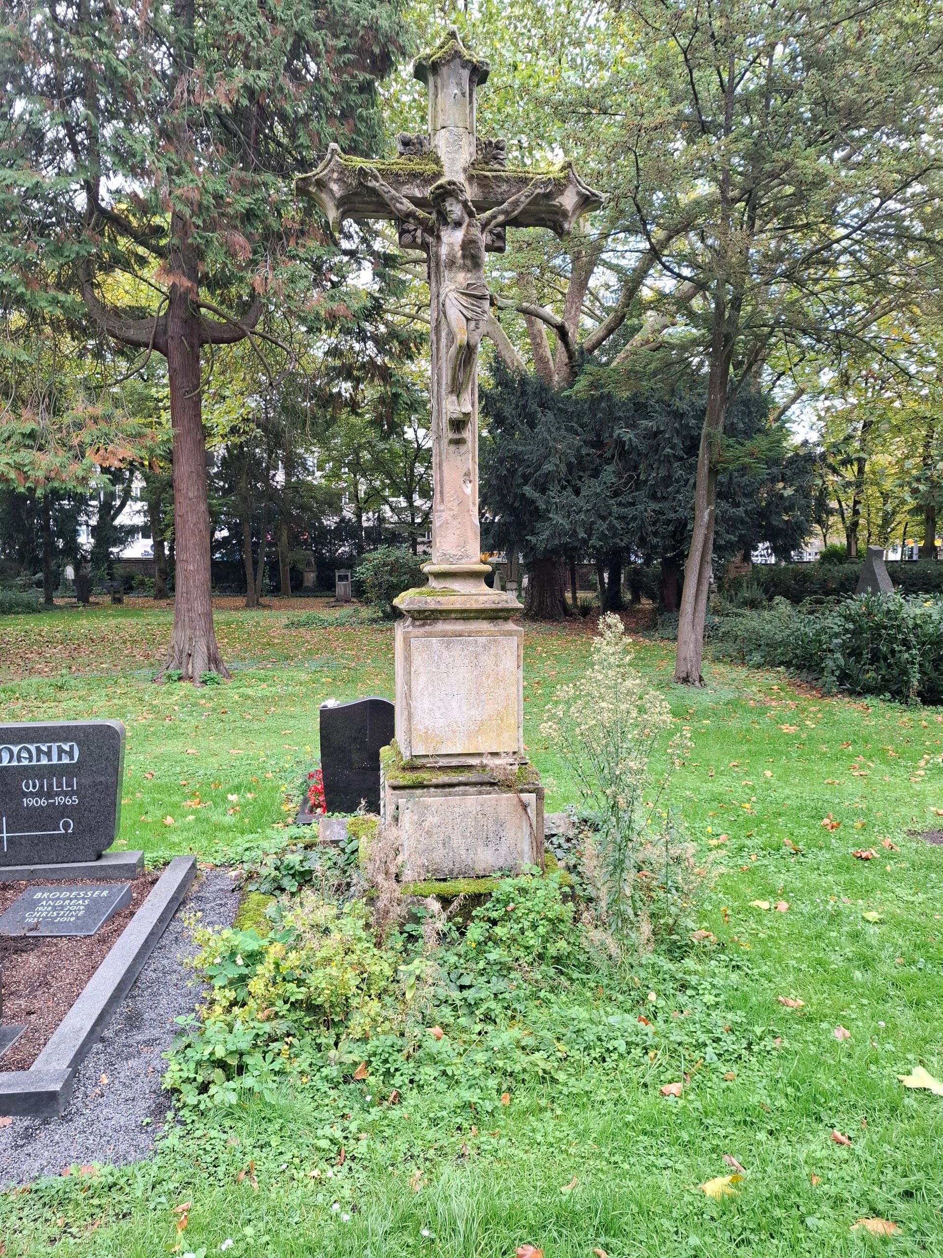 Steinkreuz auf einem Friedhof mit einer Jesusfigur. Im Hintergrund grünes Gras und Bäume.