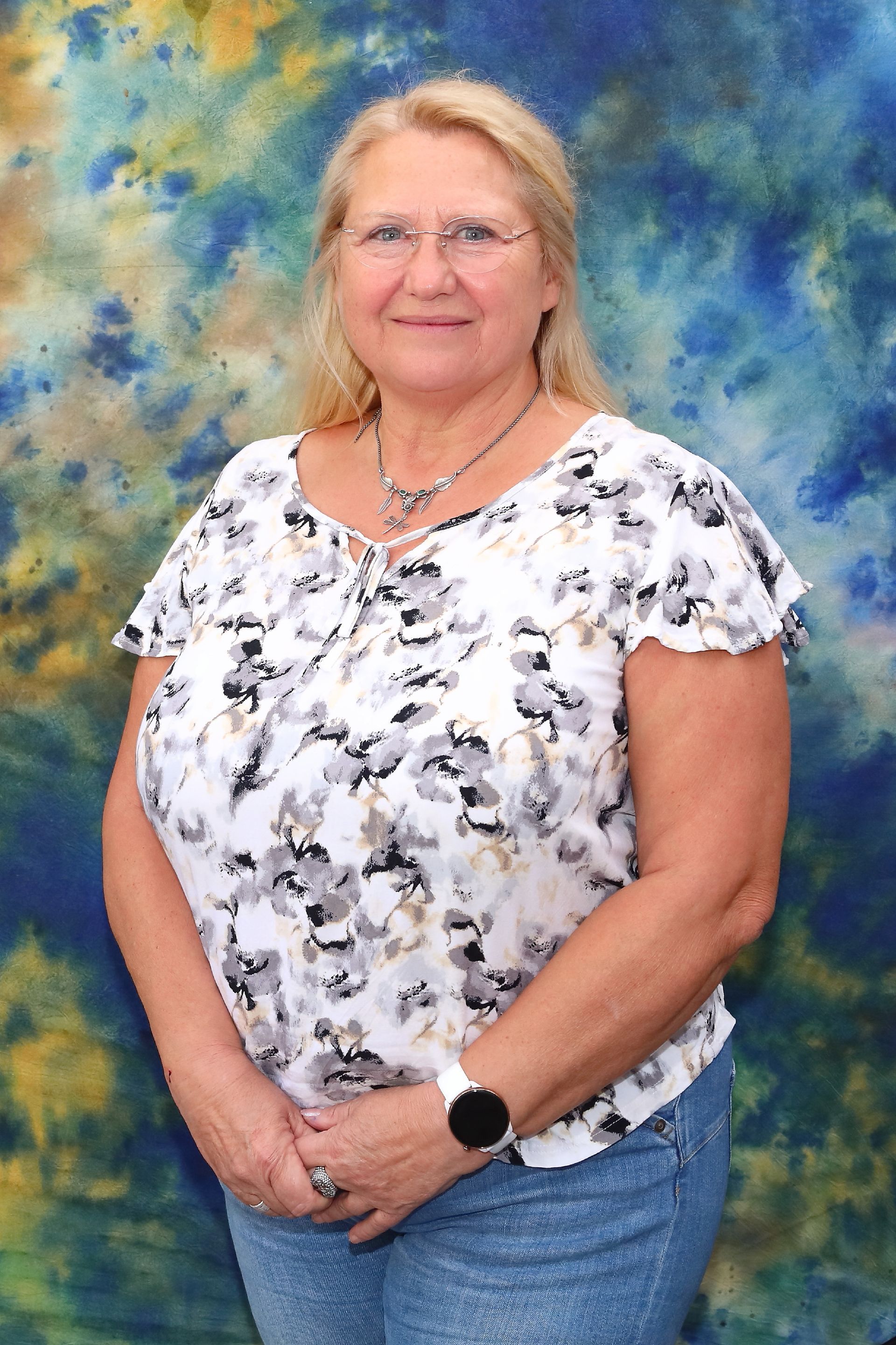 Eine Frau mit Brille, die eine geblümte Bluse und Jeans trägt, lächelt vor einem blau-gelben Hintergrund in die Kamera.