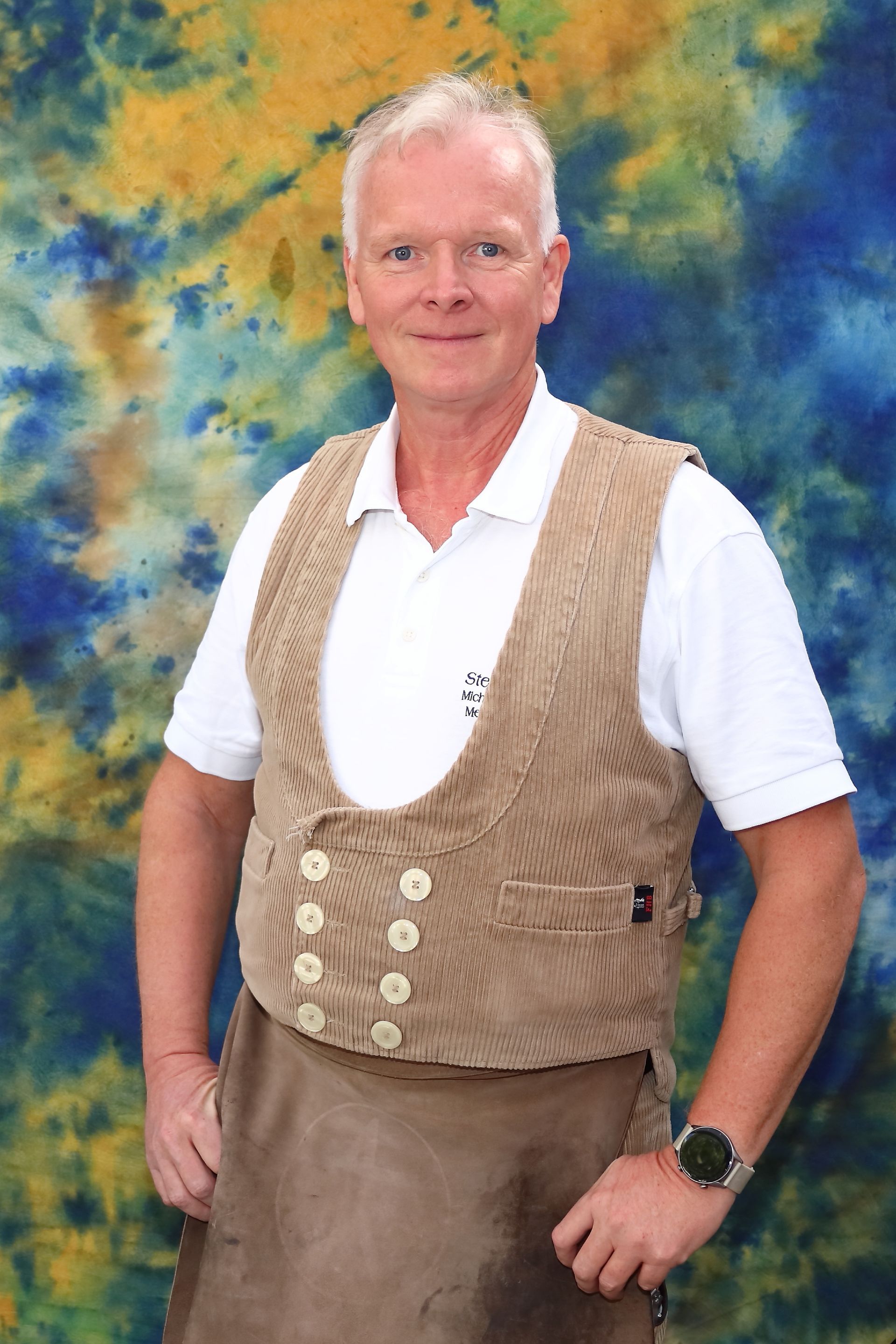 Mann in Weste und Schürze, lächelnd, vor einem farbenfrohen Hintergrund.
