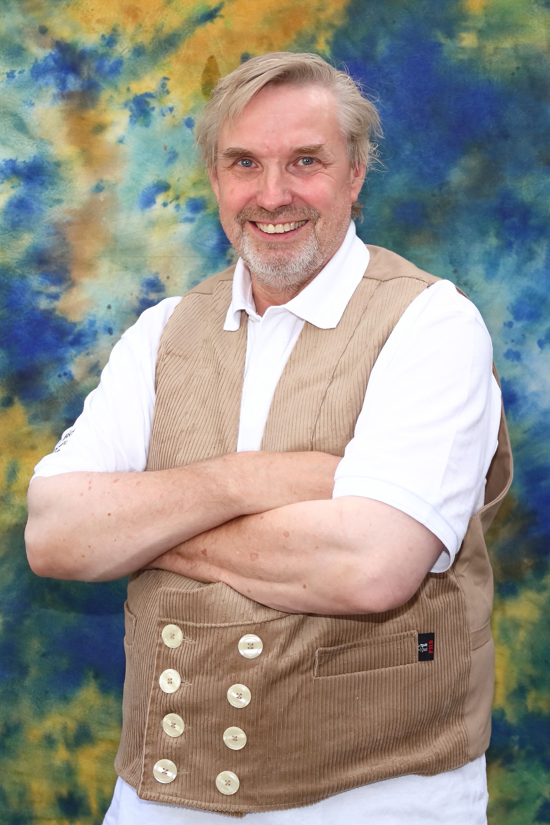 Mann in Weste mit verschränkten Armen, lächelnd, vor farbenfrohem Hintergrund.