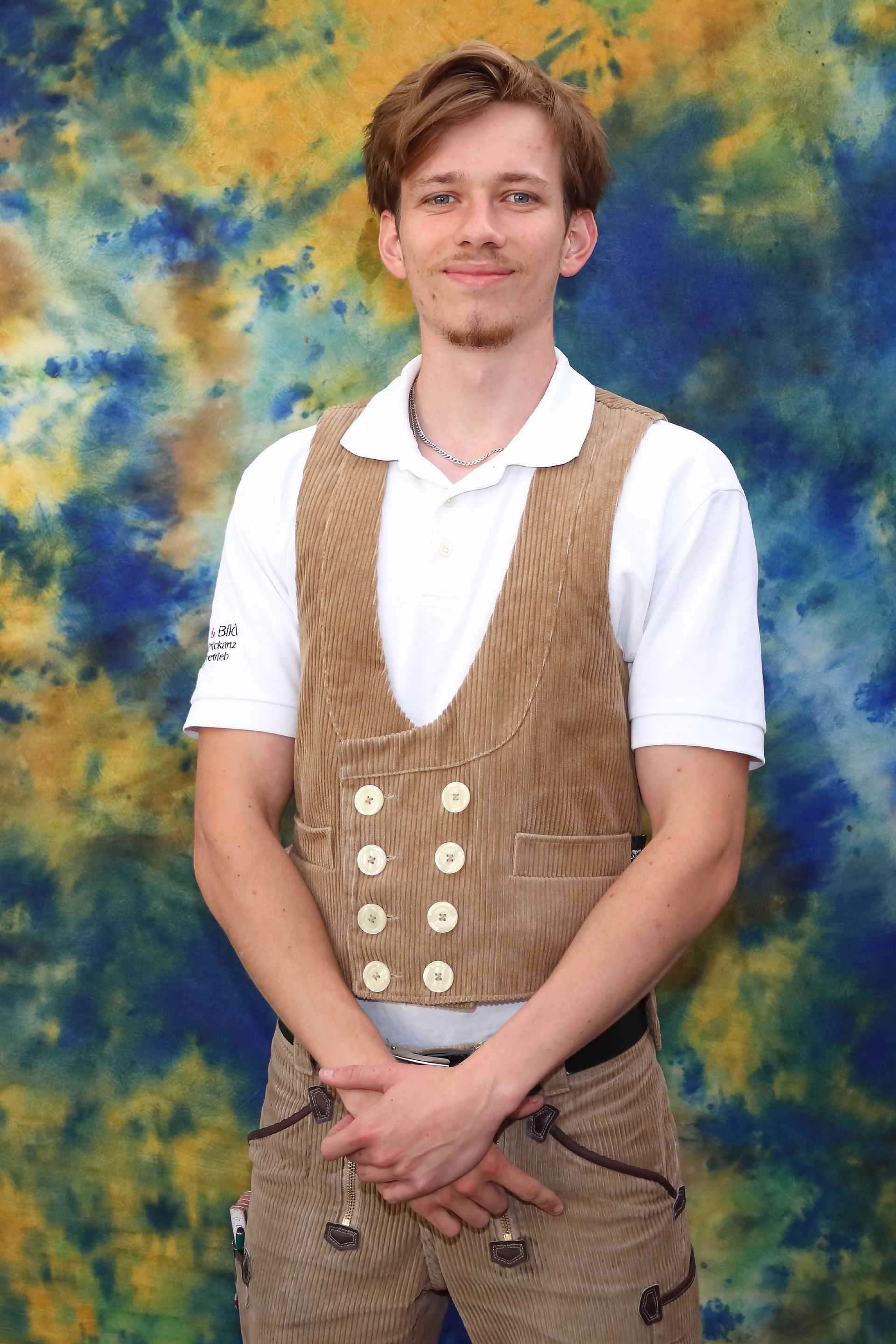 Ein Mann in traditioneller brauner Weste und Hose, weißem Hemd, die Hände gefaltet, steht vor einem farbenfrohen Hintergrund.