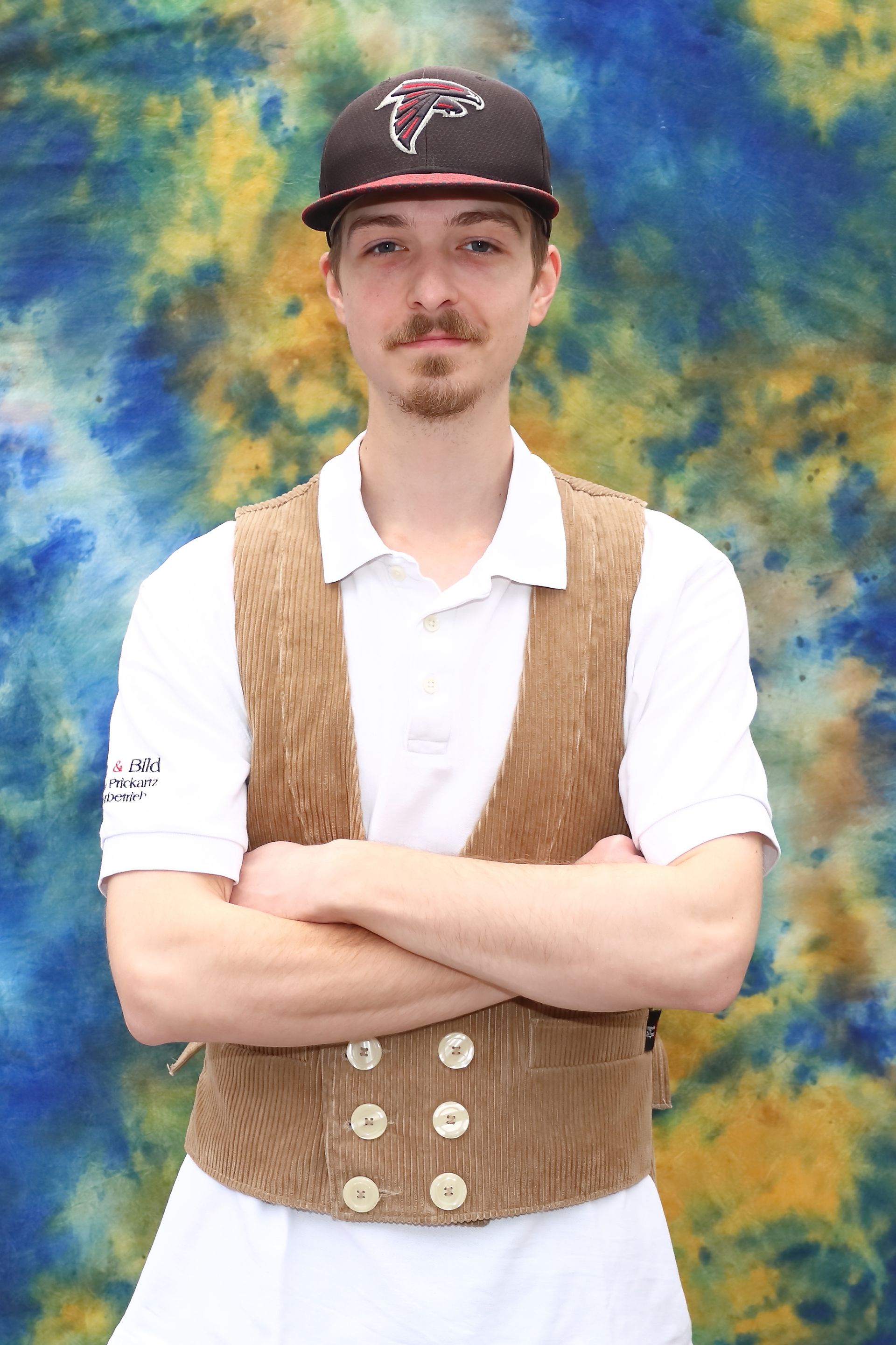 Mann in brauner Weste und Baseballkappe, die Arme verschränkt. Blau-gelber Batik-Hintergrund.